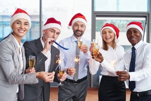 Five people in business attire wearing Santa hats celebrate indoors with champagne, sparklers, and party horns.