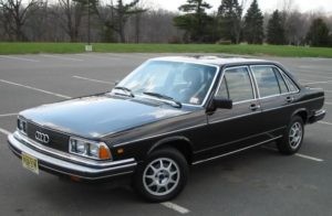 A black 1980s Audi sedan is parked in an empty outdoor parking lot with trees and grass in the background.