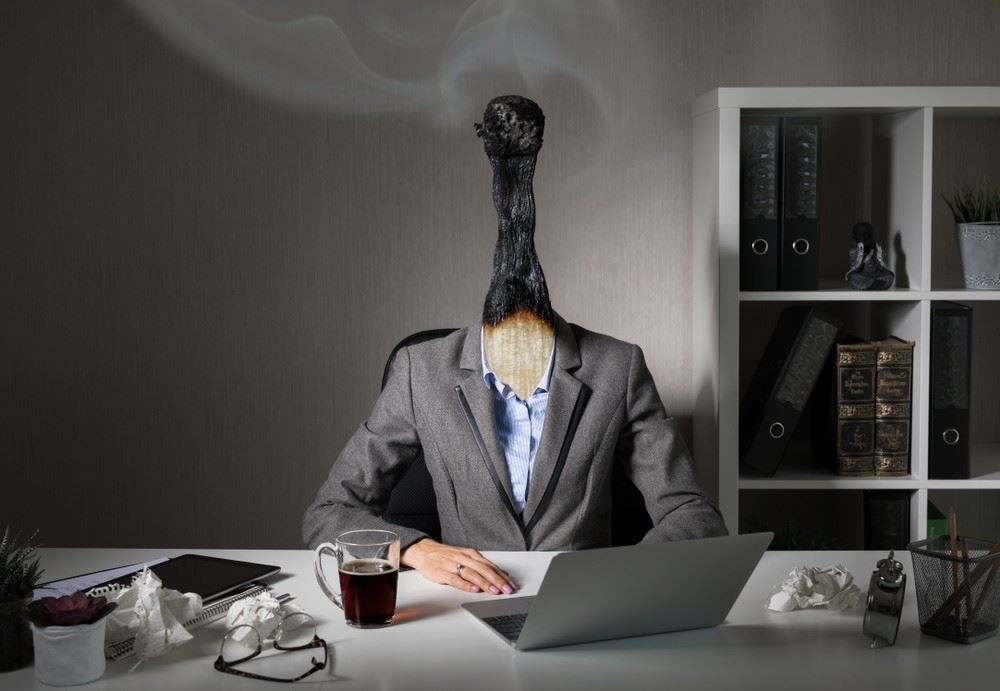 A person in a suit sits at a desk with a burned-out matchstick for a head, suggesting burnout, surrounded by office supplies and a laptop.