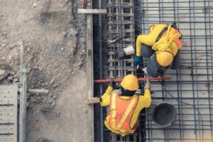 Two construction workers in safety vests and helmets are working with steel rebar and concrete at a construction site, viewed from above.