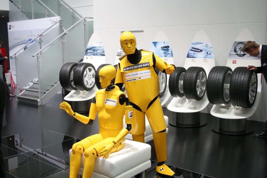Two yellow crash test dummies are posed in front of several car tires on display stands at an indoor automotive exhibition.