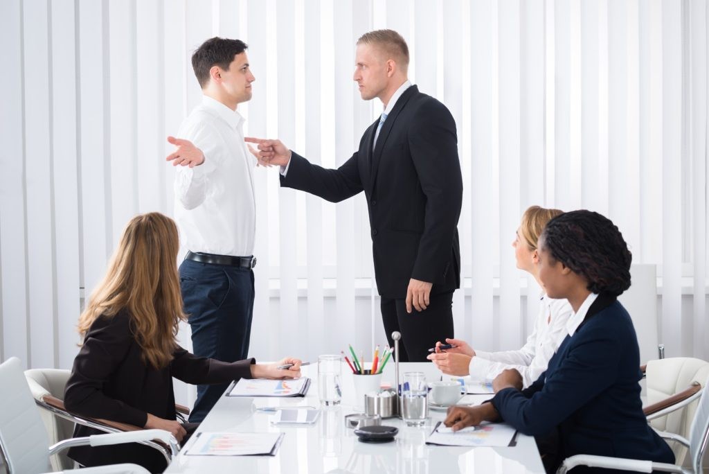 Two men standing and arguing at a conference table while three seated women observe the confrontation in a modern office setting.