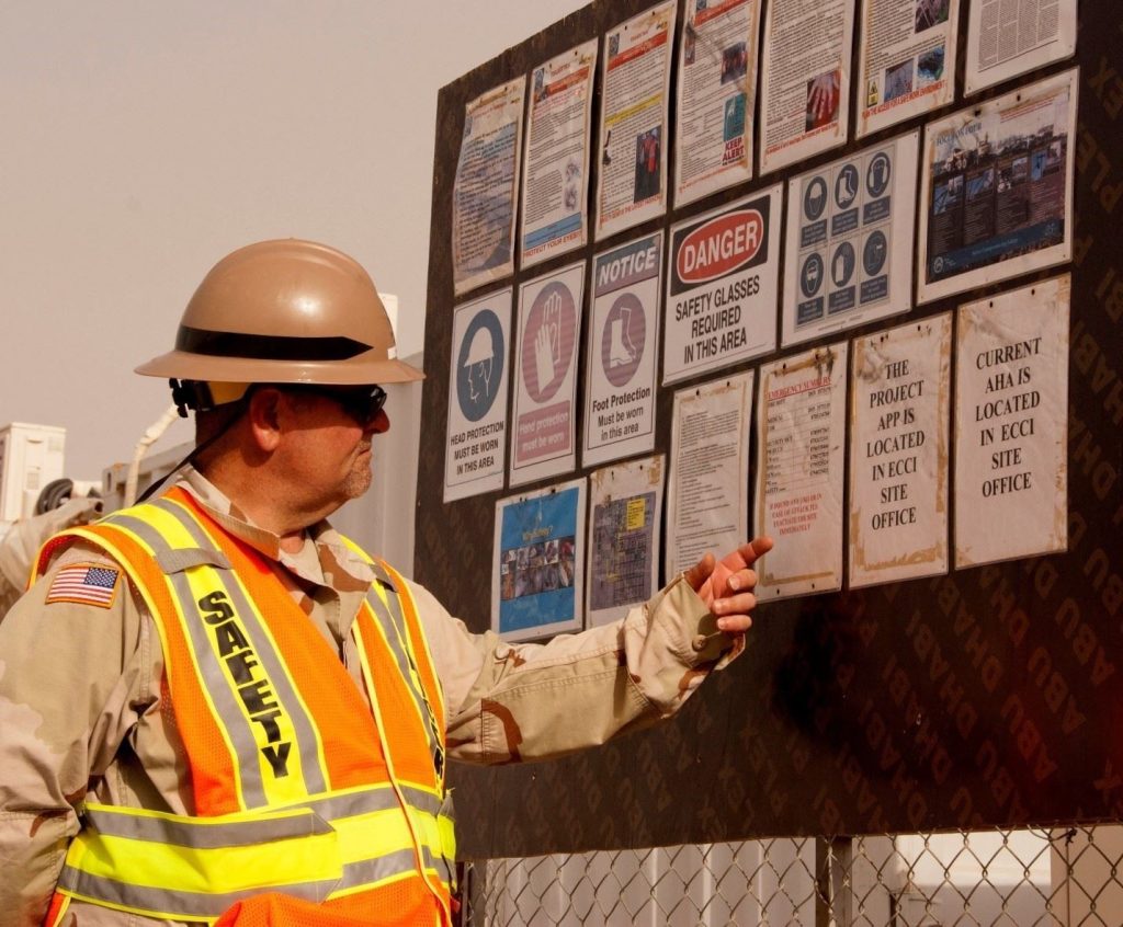A worker in safety gear points to a project information board displaying safety notices and site instructions at an outdoor construction site.