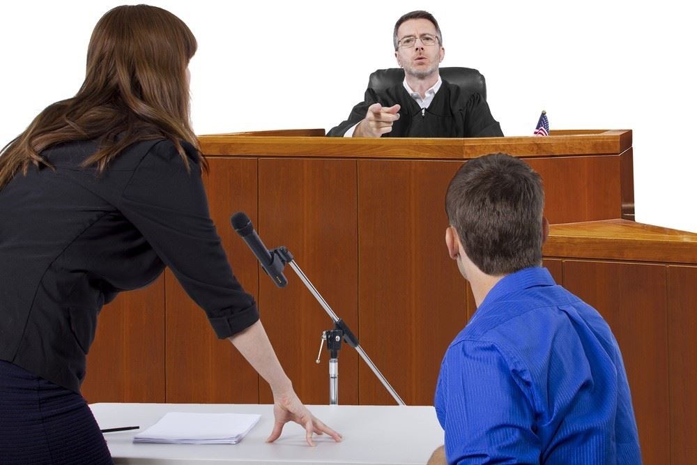 A judge points from the bench in a courtroom while a woman stands and speaks to a seated man at a table with papers and a microphone.