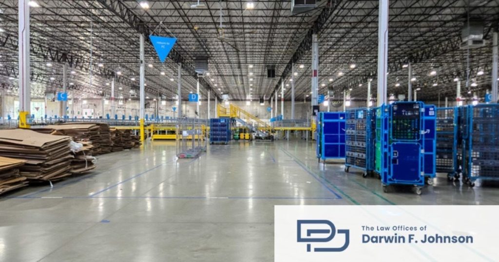 Large warehouse interior with stacks of cardboard, blue rolling carts, high ceilings, and overhead lights; logo for The Law Offices of Darwin F. Johnson in the corner.