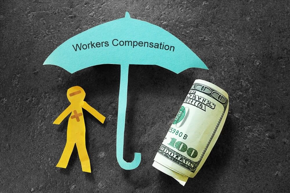 Cutout of a person and a blue umbrella labeled "Workers Compensation" next to a rolled-up $100 bill on a dark surface.