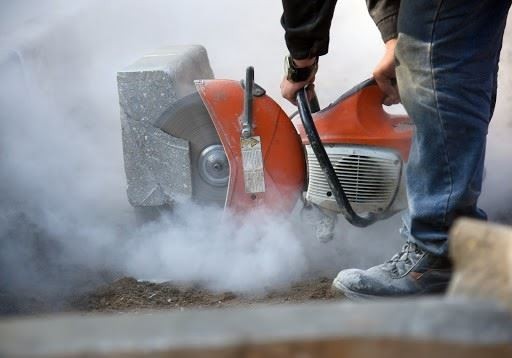 Person operating an orange power saw, cutting into concrete and creating a cloud of dust. Only lower body and tool are visible.