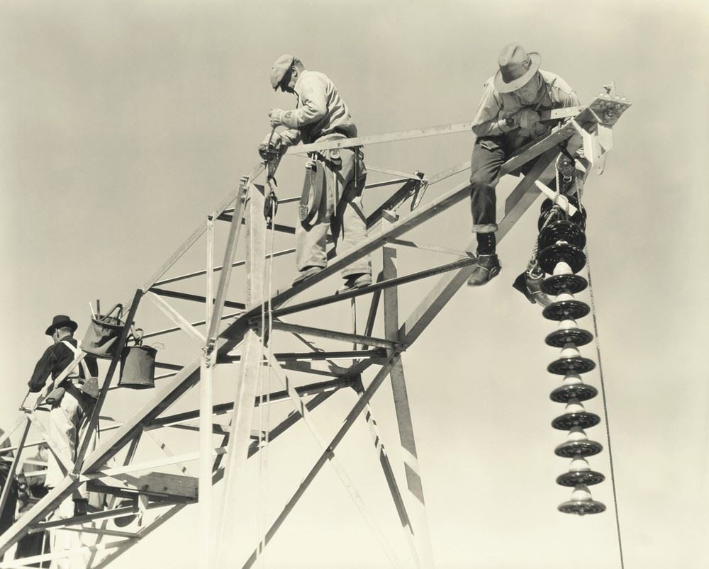 Three workers are constructing or repairing a steel electrical transmission tower, with one handling a string of large disc insulators.