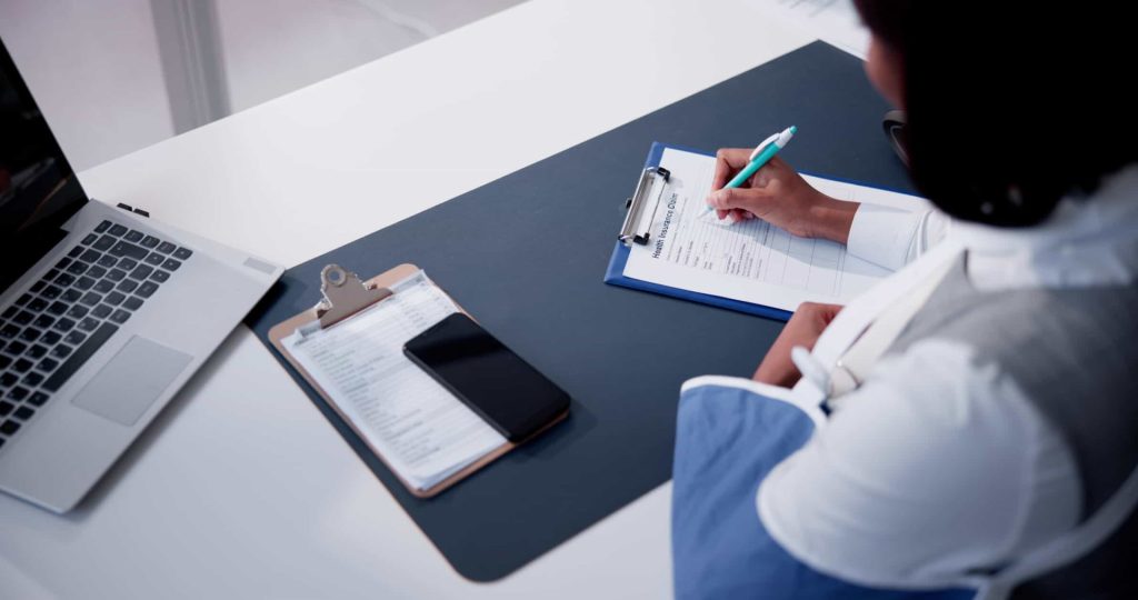Person with an arm in a sling fills out a form on a clipboard at a desk with a laptop, clipboard, and smartphone.