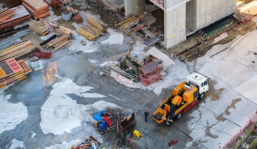 Aerial view of a construction site with scattered materials, concrete surfaces, and a white truck with yellow equipment parked near a building structure.