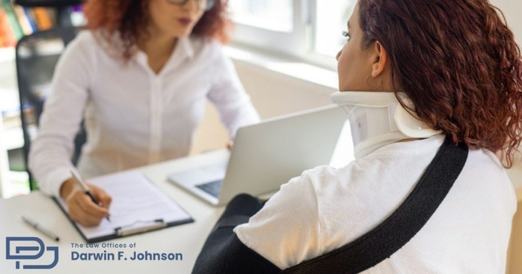 A woman with a neck brace and arm sling speaks with a professional taking notes at a desk. The logo of "The Law Offices of Darwin F. Johnson" is visible.