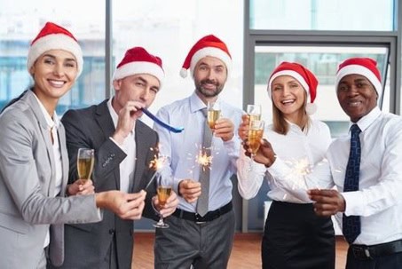 Five people wearing Santa hats celebrate indoors with sparklers, champagne, and party favors, smiling at the camera.
