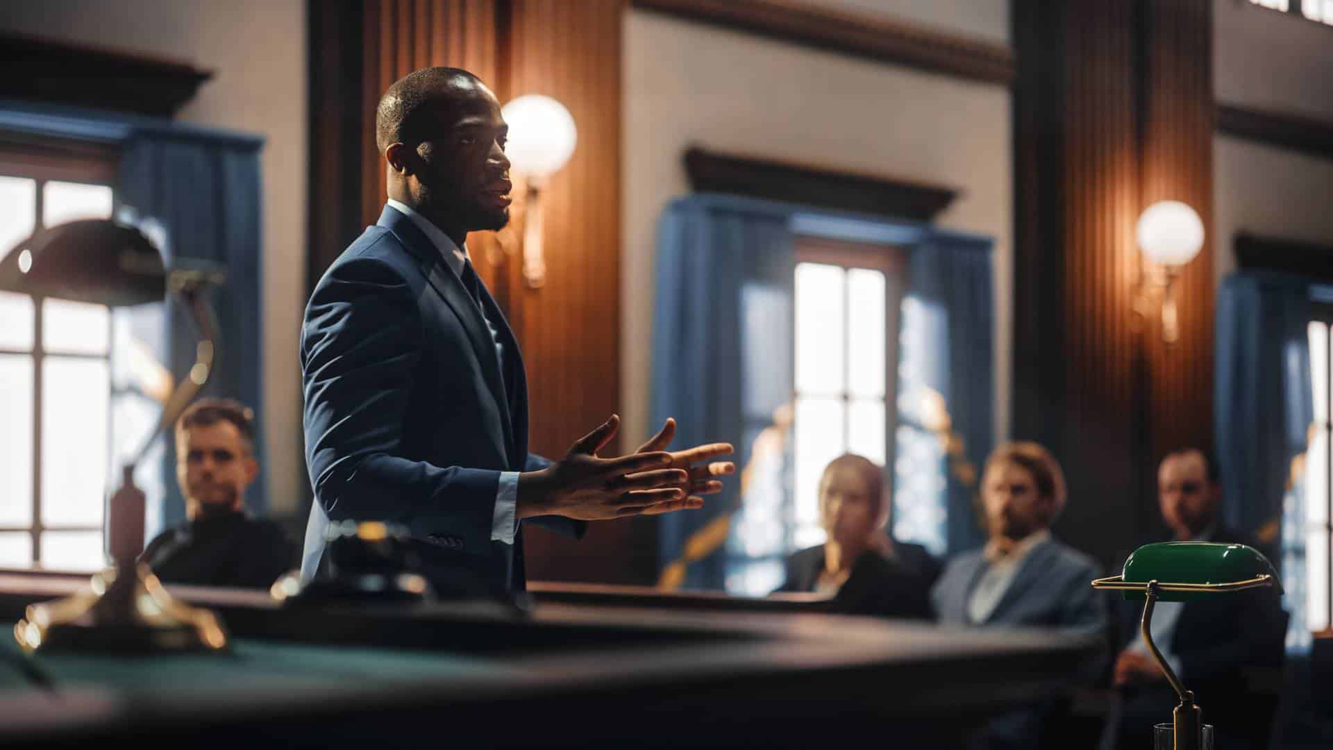 A man in a suit speaks to a seated audience in a formal room with wooden paneling and large windows.