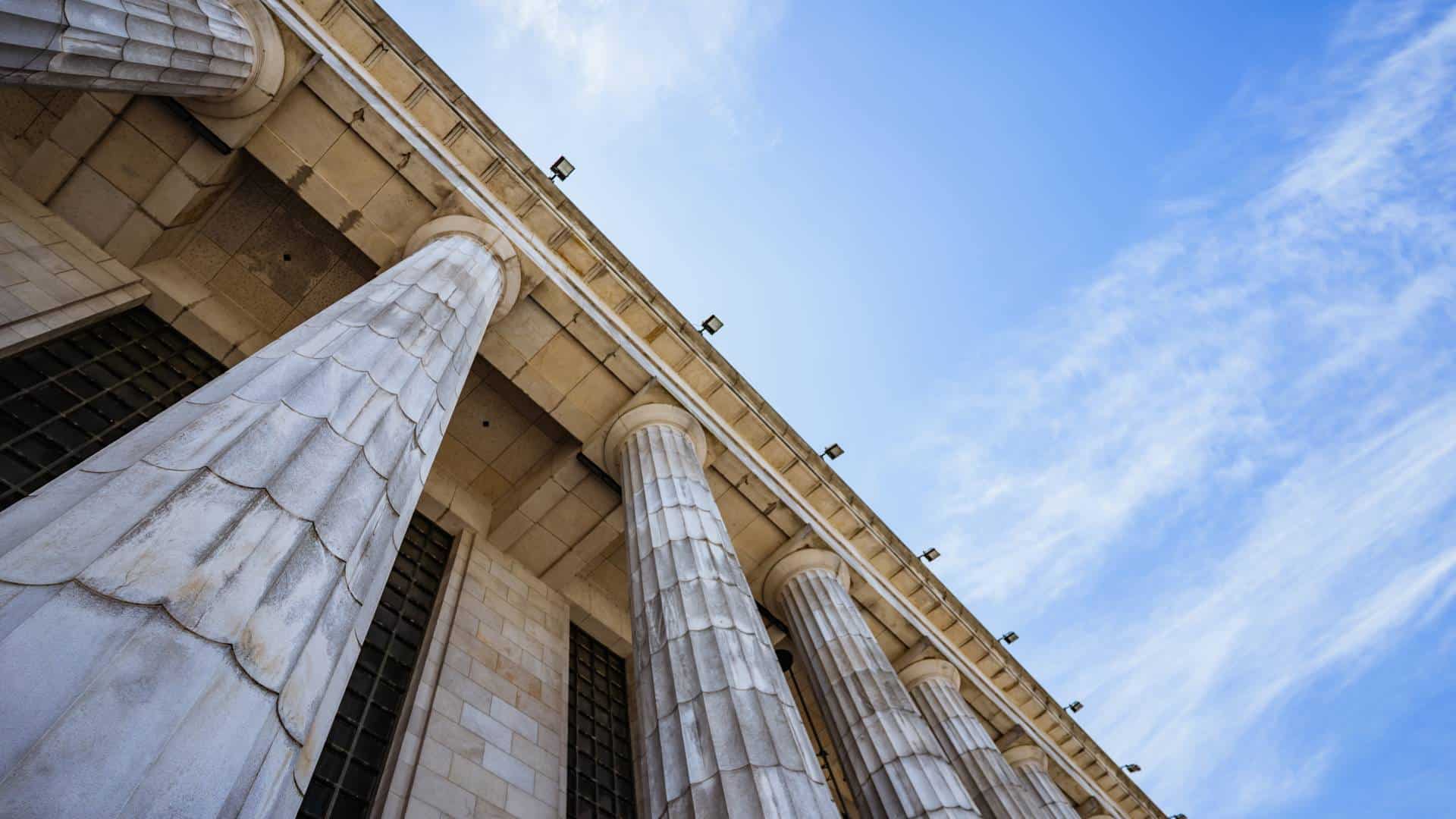 Low-angle view of large stone columns supporting a neoclassical building against a blue sky with wispy clouds.