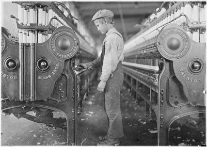 A young boy wearing work clothes and a cap stands between rows of large textile machines in a factory, operating or inspecting the equipment.