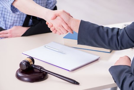 Two people shaking hands across a desk with a gavel, notebook, pen, and book visible on the tabletop.