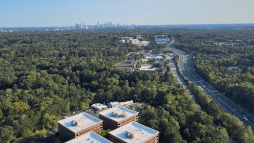 Aerial view of a highway running through a forested area with office buildings in the foreground and a distant city skyline on the horizon.