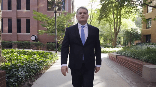 A man in a dark suit and blue tie walks confidently down a tree-lined sidewalk between brick buildings on a sunny day.