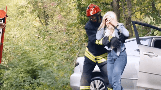 A firefighter helps a distressed woman away from a silver car with an open door in a wooded area.
