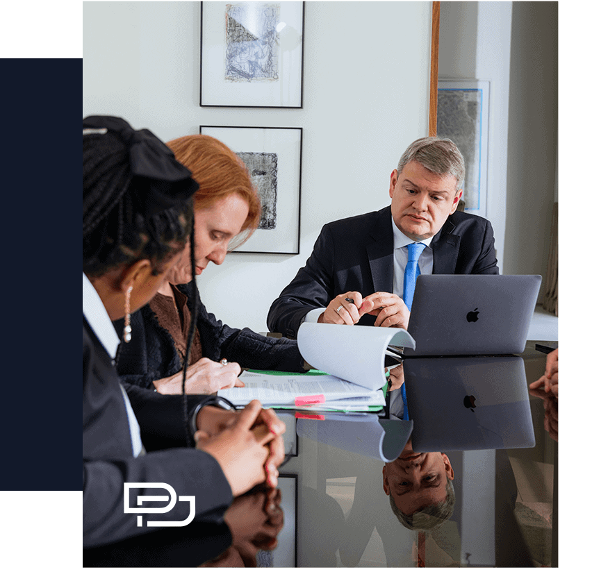 Three people in business attire sit at a conference table with documents and a laptop, appearing focused during a meeting.