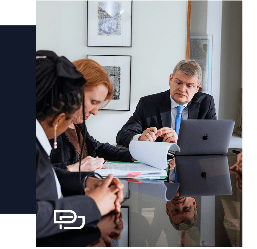 Three people in business attire sit at a table with documents and a laptop, engaged in a meeting. A man in a suit listens intently, while two women review papers.