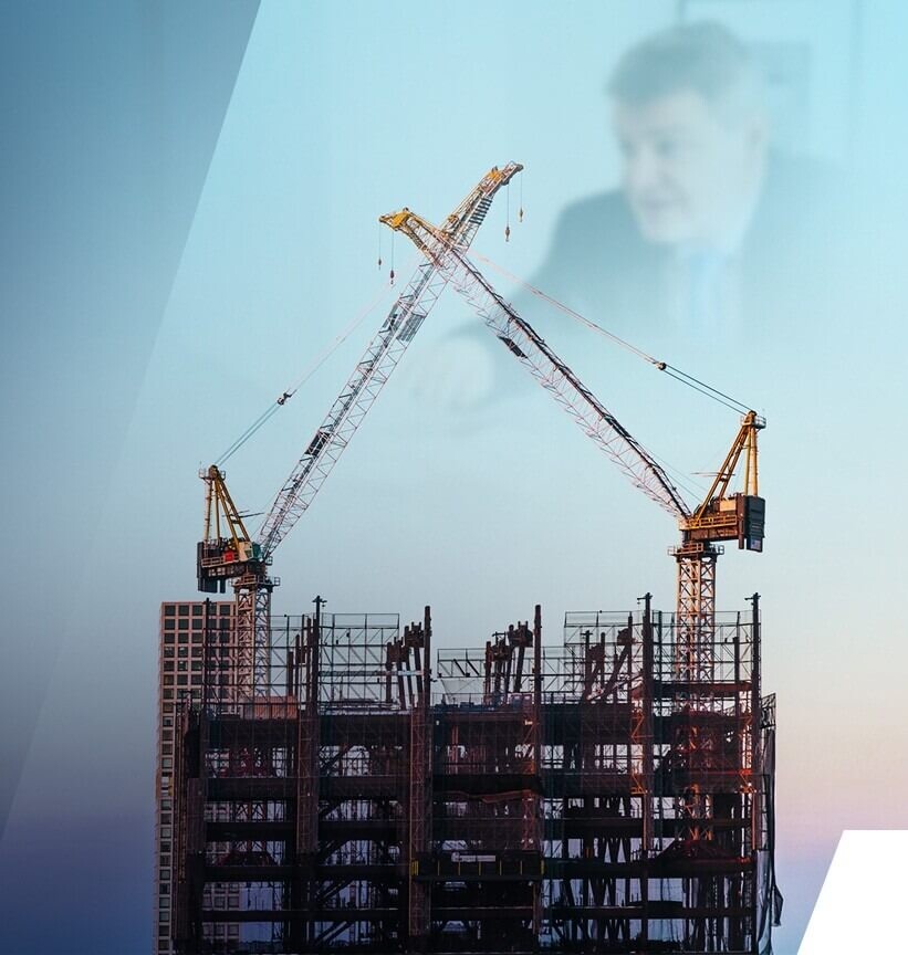 Two construction cranes work on a high-rise building frame, with the blurred image of a man in a suit visible in the background.