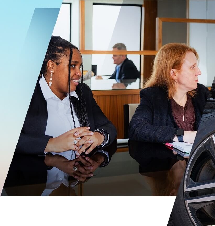 Two women in business attire sit at a conference table, engaged in conversation, with two more people in the background.