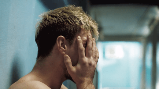 A shirtless person with blond hair stands against a blue wall, covering their face with one hand.