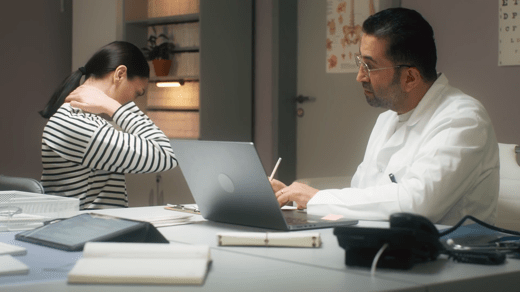 A woman holds her neck in discomfort while sitting across from a doctor who is taking notes at his desk.