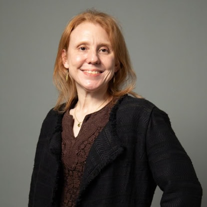 A woman with light brown hair wearing a dark jacket and brown blouse stands against a plain gray background, smiling at the camera.