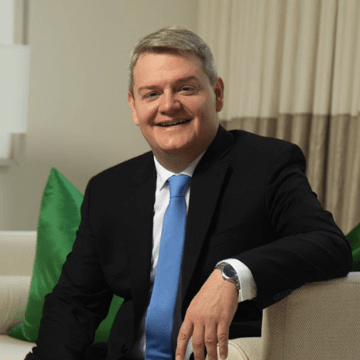 A man in a dark suit and light blue tie sits on a light-colored sofa with green cushions, smiling at the camera.