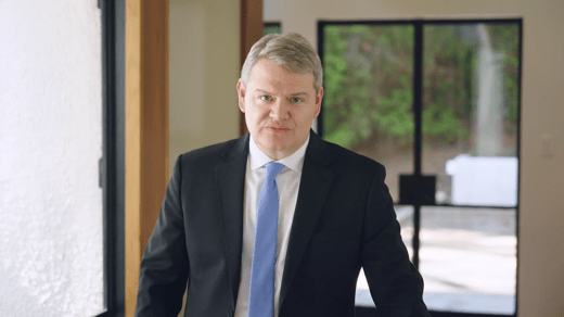 A man in a suit and blue tie stands indoors near a glass door, looking directly at the camera.