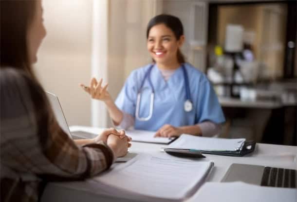 A healthcare professional in scrubs speaks with a patient across a desk with documents, a clipboard, and a stethoscope visible.