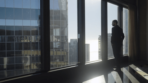 A man in a suit stands by large windows in a high-rise office, looking out at city buildings in daylight.