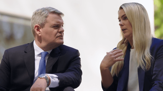 Two people in business attire sit and talk outdoors; the man listens while the woman gestures with her hand as she speaks.