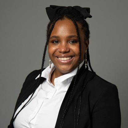 A woman with long braids and a black bow in her hair smiles, wearing a black blazer and white shirt against a plain gray background.