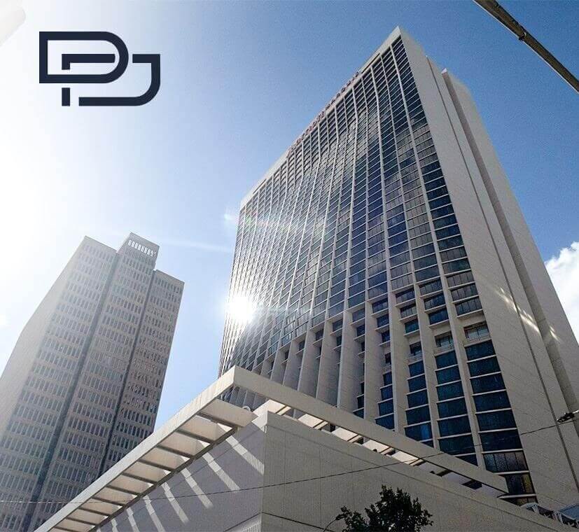 Two modern high-rise office buildings against a blue sky, viewed from below, with sunlight reflecting off the glass. A minimalist "PJ" logo is present in the upper left corner.