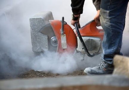 A person using an orange power saw to cut concrete, creating a cloud of dust around the work area.