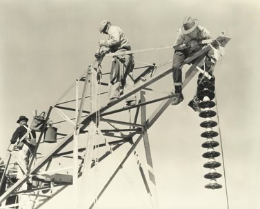 Three workers in hats and protective clothing assemble or maintain a high-voltage transmission tower, handling tools and large insulator discs against a clear sky.