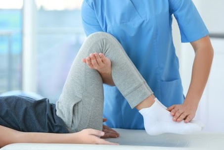 A person in grey pants receives physical therapy as a healthcare professional in blue scrubs supports their bent leg and ankle.
