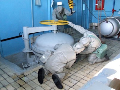 Three workers in protective suits operate a large yellow valve in an industrial facility with tiled flooring and blue walls.
