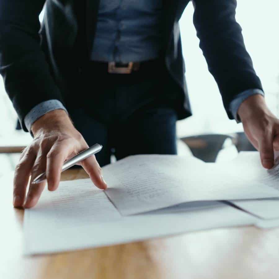 Person in business attire leaning over a desk, holding a pen and reviewing documents spread out on the table.