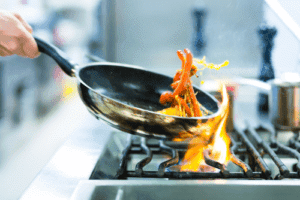 A person cooks sliced vegetables in a frying pan over a gas stove with visible flames.