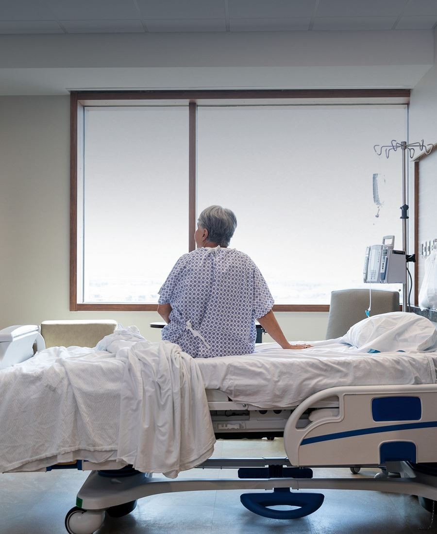 An older patient in a hospital gown sits on a bed facing a window, with medical equipment and an IV stand nearby.