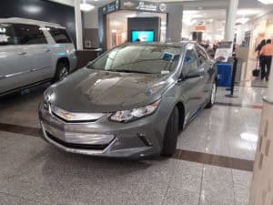 A gray Chevrolet sedan is displayed inside a shopping mall, positioned on a tiled floor near storefronts and other vehicles.