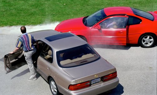 A man exits a beige car after a collision with a red car on a paved road. Both vehicles show damage and are surrounded by smoke or dust.