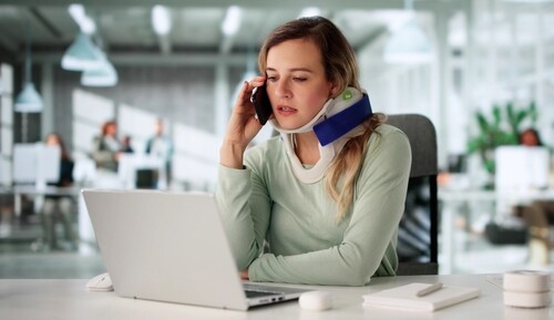 woman wearing cervical neck brace talking on phone at office desk with laptop