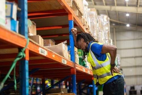 warehouse worker in safety vest clutching back in pain near storage shelves